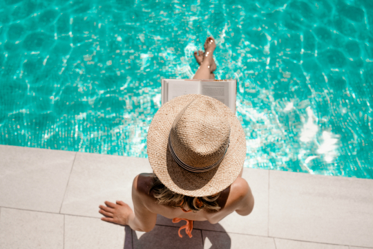Frau mit Strohhut von oben, sitzt an Poolrand mit den Beinen im Wasser und liest ein Buch
